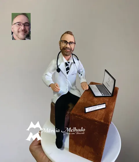 Boneco de biscuit de médico barbudo com óculos, jaleco branco e estetoscópio, sentado à mesa de madeira com notebook aberto