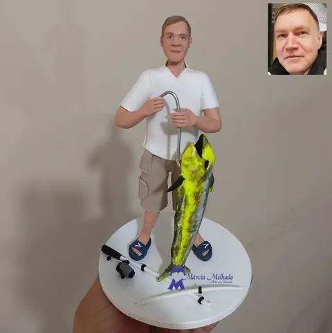 Boneco de biscuit de pescador com camisa polo branca e bermuda segurando anzol, ao lado de um peixe dourado grande