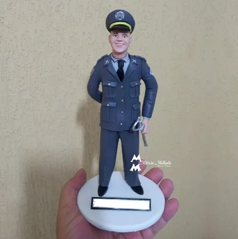 Boneco de biscuit de policial militar em uniforme de gala cinza com quepe e espada, sobre base branca com plaquinha