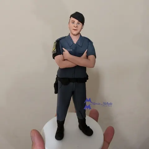 Boneco de biscuit de policial militar com farda cinza e boina preta, de braços cruzados sobre base branca