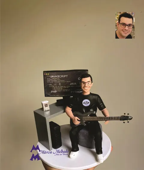 Boneco de biscuit de óculos e camiseta preta tocando baixo sentado diante de mesa com monitor de códigos de programação