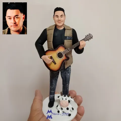 Boneco de biscuit de rapaz tocando violão, com camiseta preta, colete bege e calça jeans, e uma vaquinha malhada aos pés
