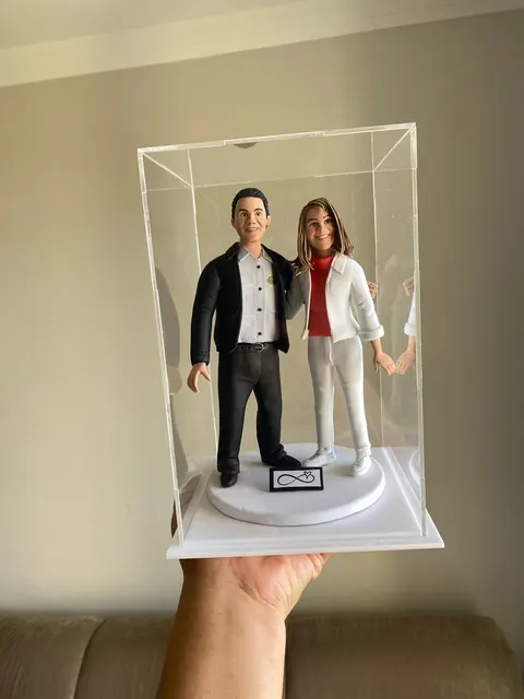 Casal de bonecos de biscuit abraçados em caixa de acrílico, ele de blazer preto e ela de blazer branco com blusa vermelha