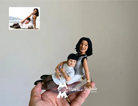 Bonecos de biscuit de mulher sentada de regata cinza e calça branca com um menino deitado no colo, sobre base transparente