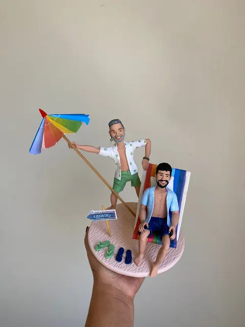 Noivinhos de biscuit na praia: um homem grisalho segurando guarda-sol colorido e outro sentado em cadeira de praia listrada