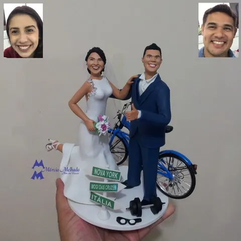 Noivos de biscuit com bicicleta azul, haltere, óculos e placa de direções indicando Nova York, Mogi das Cruzes e Itália