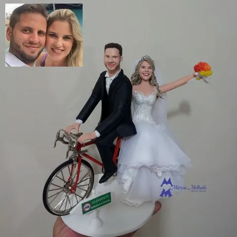 Noivo de terno preto pedalando bicicleta vermelha com a noiva de véu na garupa erguendo um buquê laranja, placa verde na base