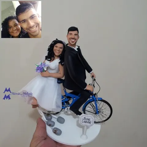 Topo de bolo de biscuit com noivo pedalando bicicleta azul, noiva na garupa com buquê lilás e placa Recém Casados