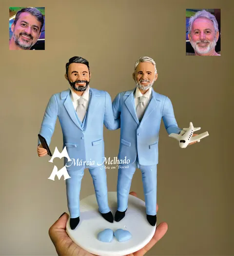 Dois noivos de biscuit em ternos azul-claros e gravatas prateadas, abraçados, um com celular e o outro com um avião