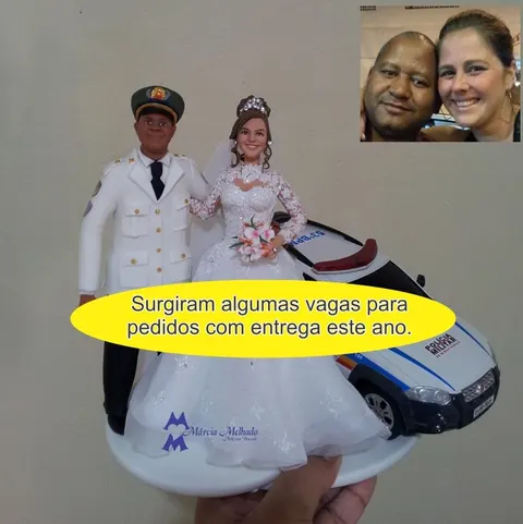 Topo de bolo de biscuit com noivo de farda branca de gala da Polícia Militar e noiva de renda ao lado de uma viatura